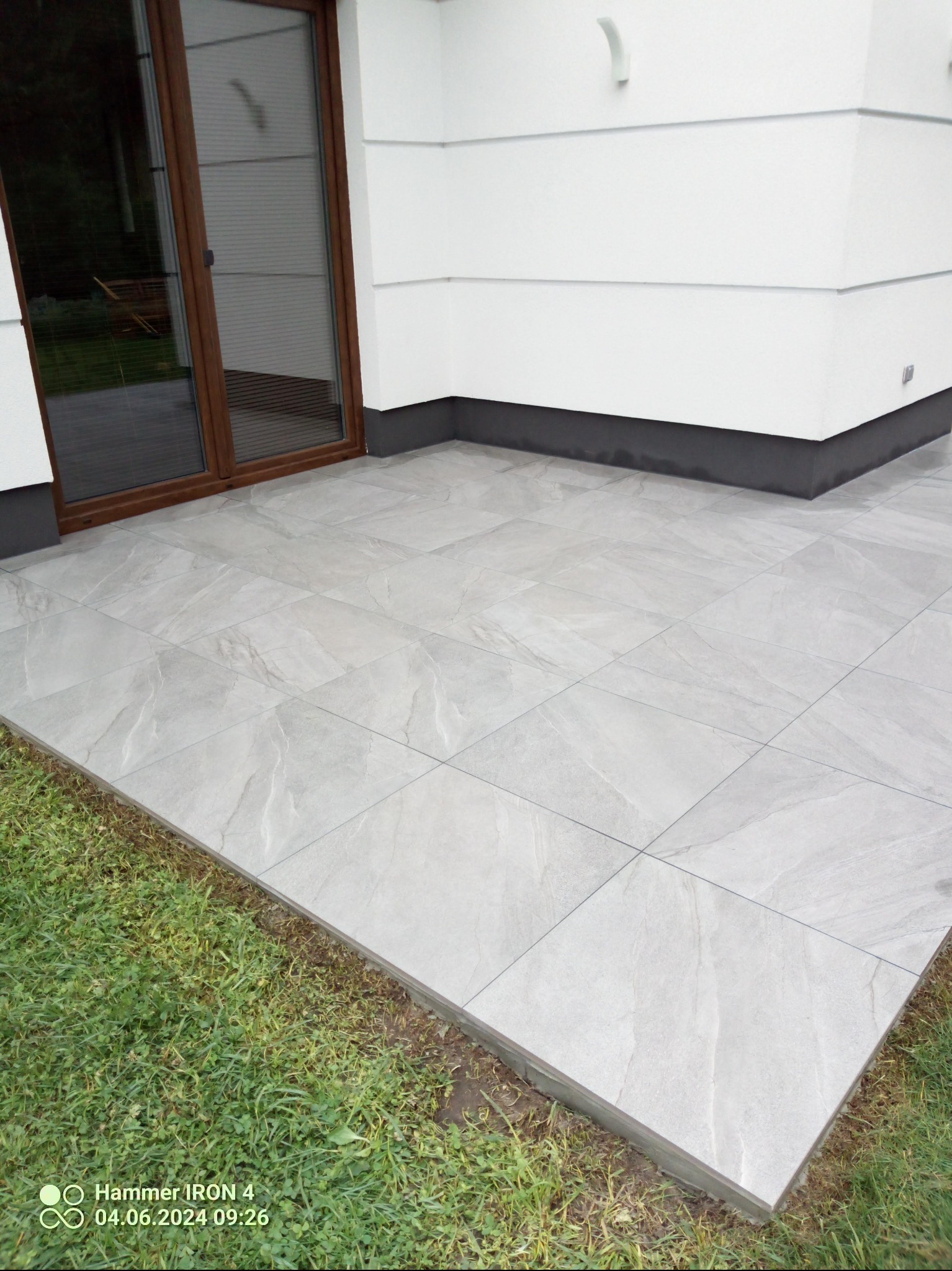 Jasnoszary taras z płytek ceramicznych, widok z góry, z brązowymi drzwiami balkonowymi i fragmentem trawnika. Minimalistyczny design.