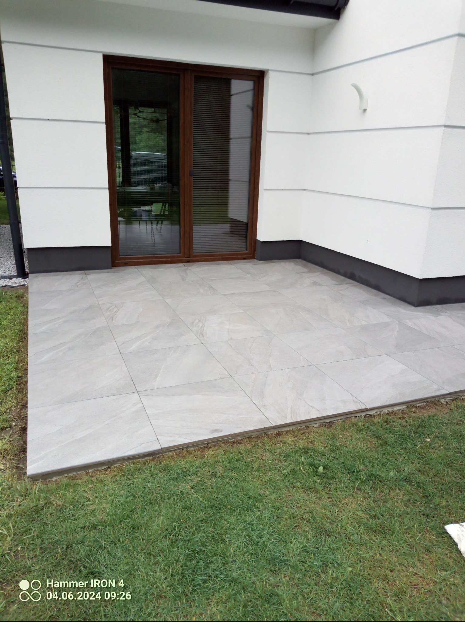 Jasnoszary taras z płytek ceramicznych przy białym budynku z brązowymi drzwiami przesuwnymi. Widok na trawnik. Minimalistyczny design.