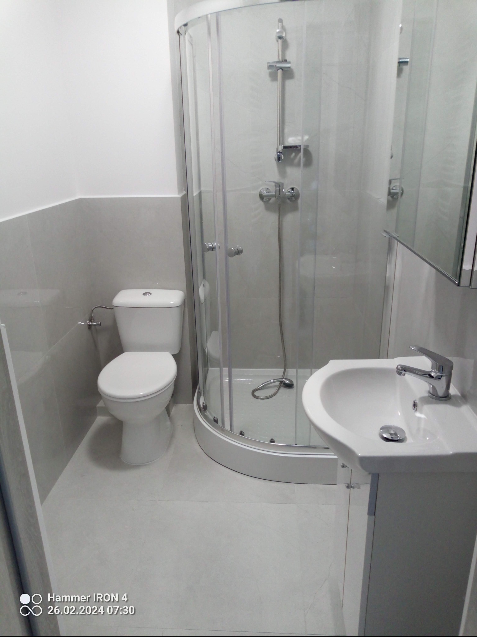 Jasna łazienka po remoncie: kabina prysznicowa, umywalka z szafką i toaleta. Ściany i podłoga wyłożone szarymi płytkami. Minimalistyczny styl, czyste linie.