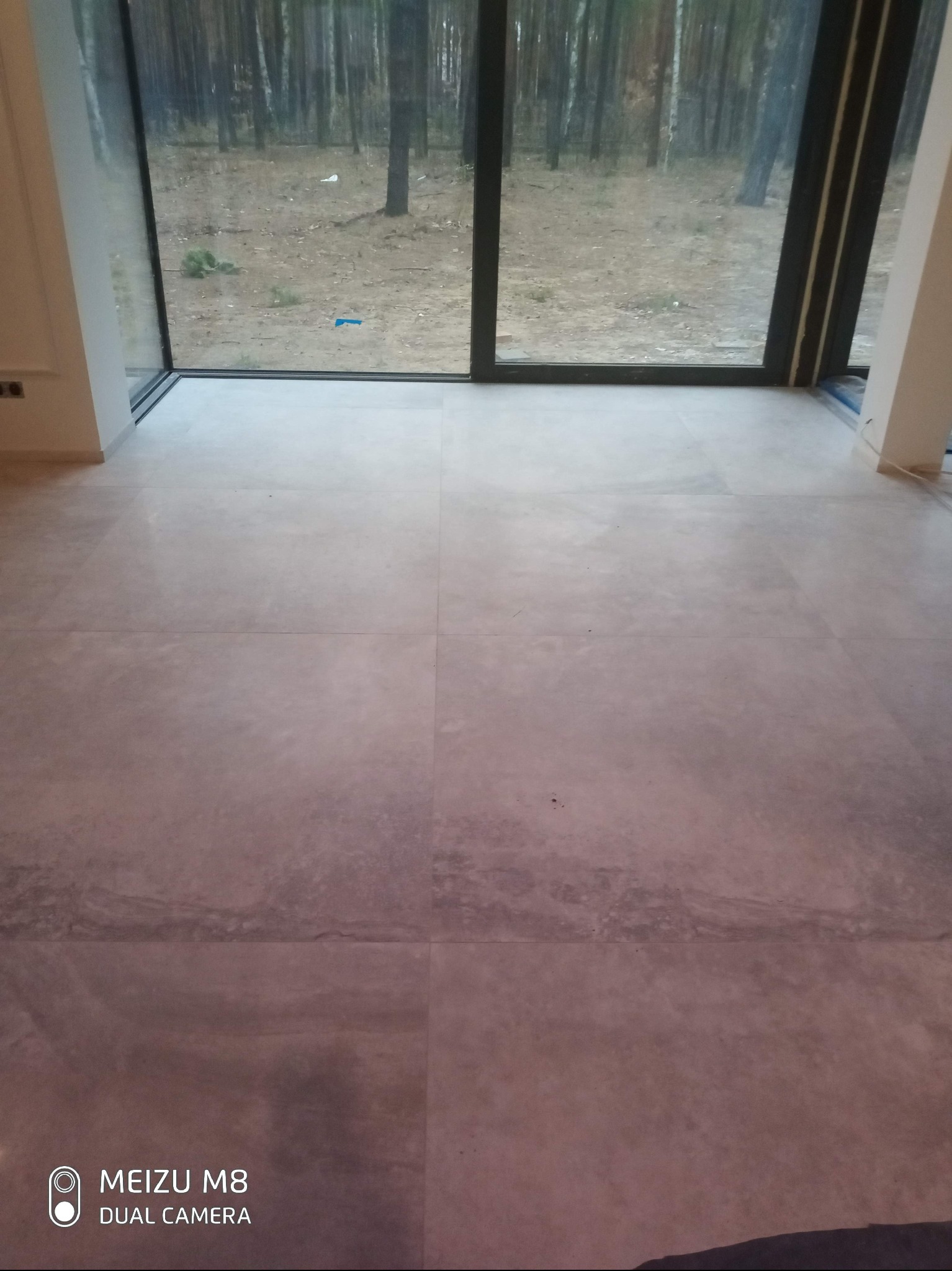 Jasne, matowe płytki podłogowe w nowym wnętrzu z dużym oknem na las. Minimalistyczny design, widoczne fugi, naturalne światło.