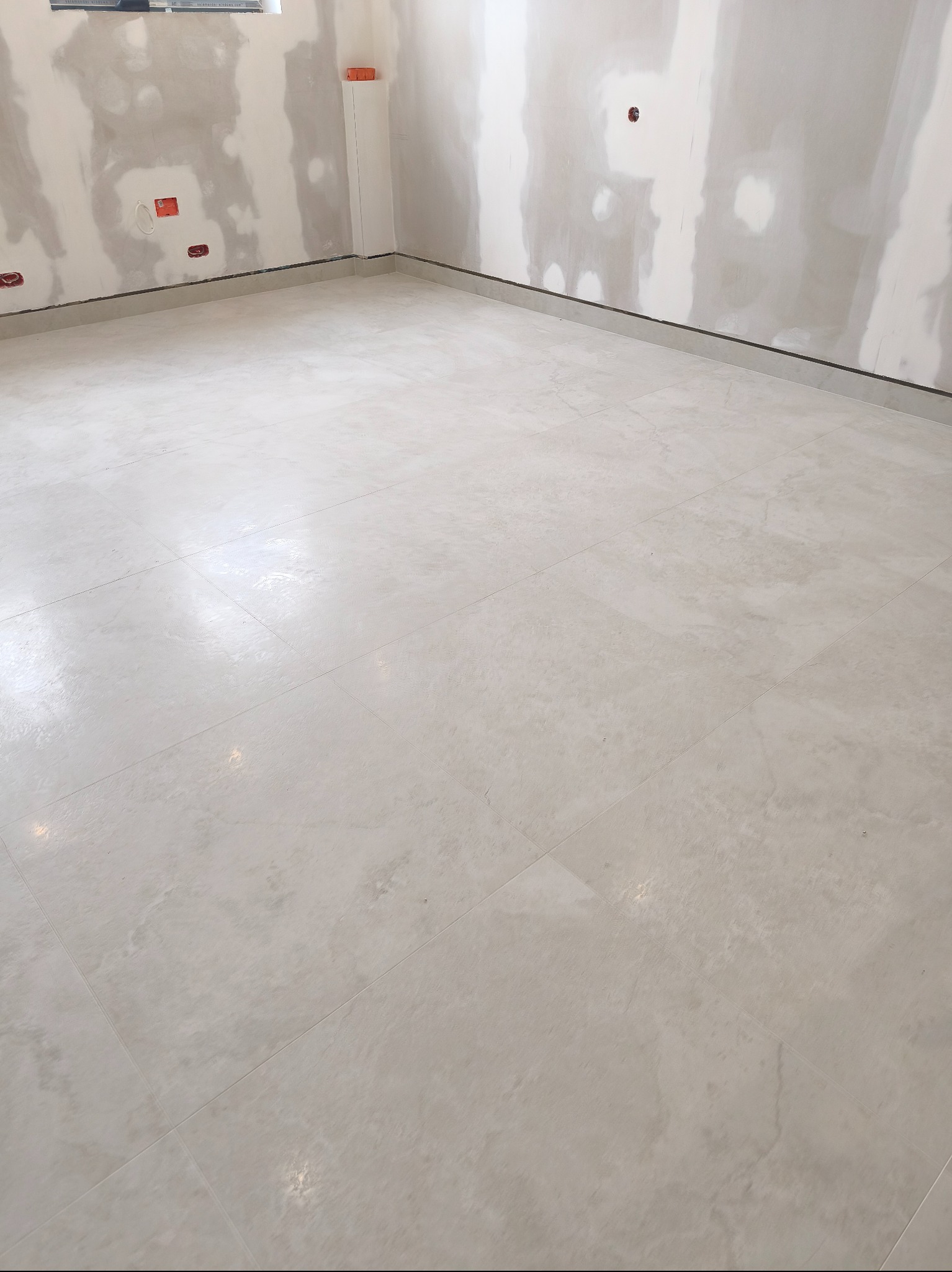 Jasne, kwadratowe płytki podłogowe w surowym wnętrzu z przygotowanymi ścianami do malowania. Widoczne gniazdka elektryczne i listwa przypodłogowa.