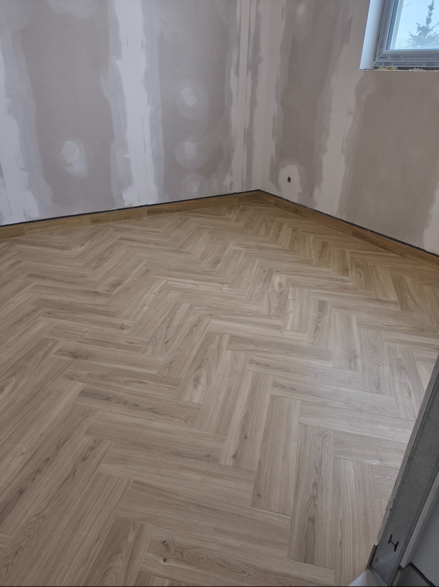Ukończona podłoga z paneli ułożonych w jodełkę w rogu pokoju. Ściany przygotowane do malowania, widoczne szpachlowane miejsca. Okno w tle.