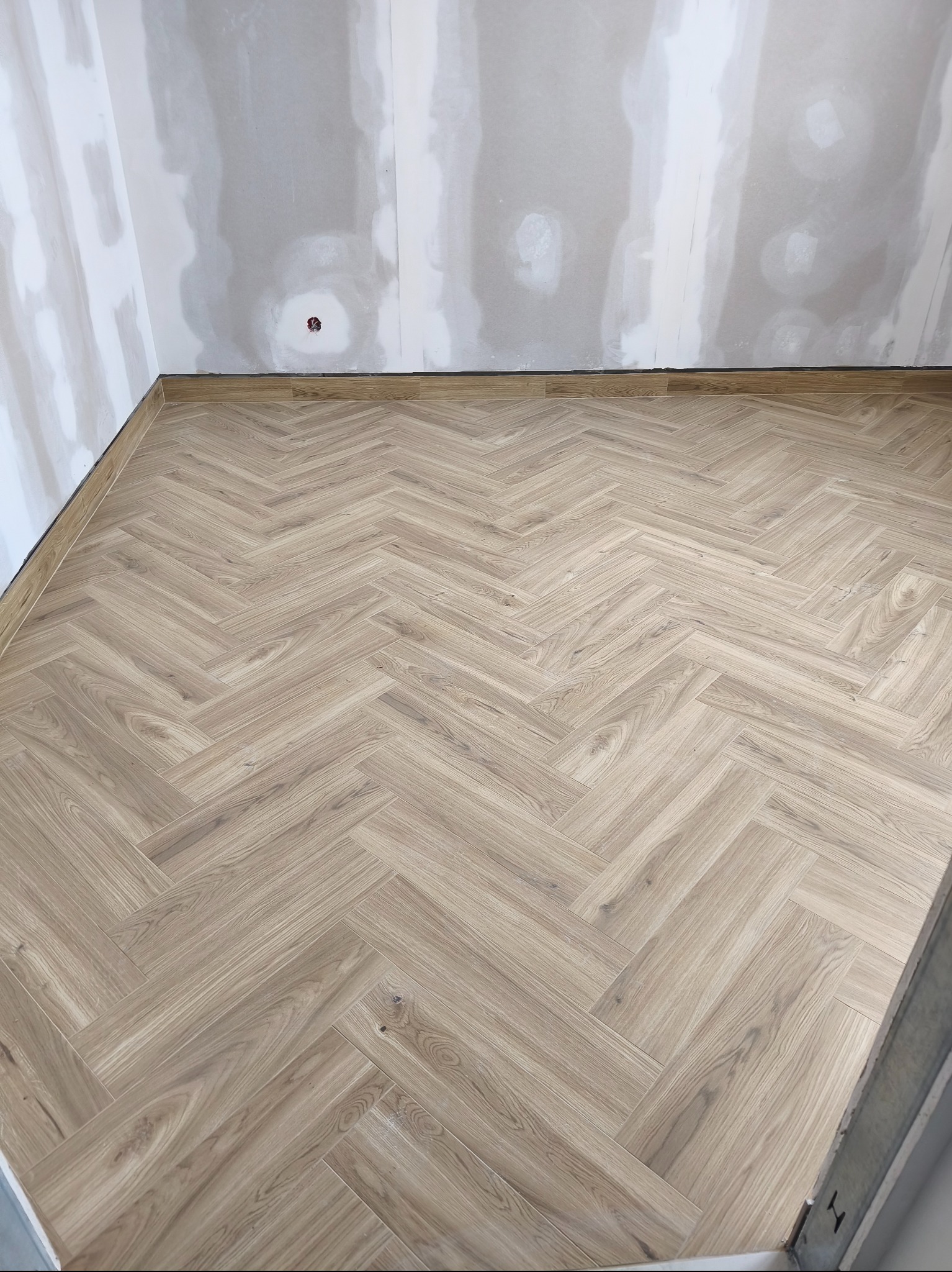 Ułożona podłoga z paneli w jodełkę w jasnym odcieniu drewna, z widocznymi surowymi ścianami przygotowanymi do dalszych prac wykończeniowych. Listwa przypodłogowa w kolorze podłogi.