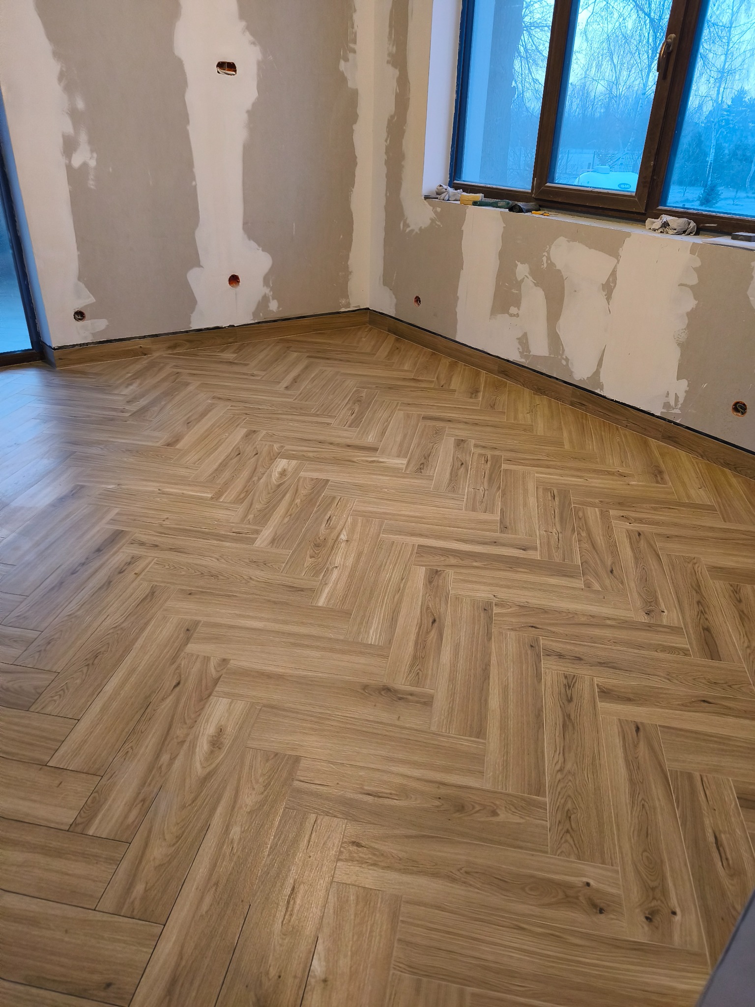 Ukończona podłoga z płytek imitujących drewno, ułożonych w jodełkę, w pomieszczeniu z częściowo wykończonymi ścianami i oknem. Widoczne gniazdka elektryczne.