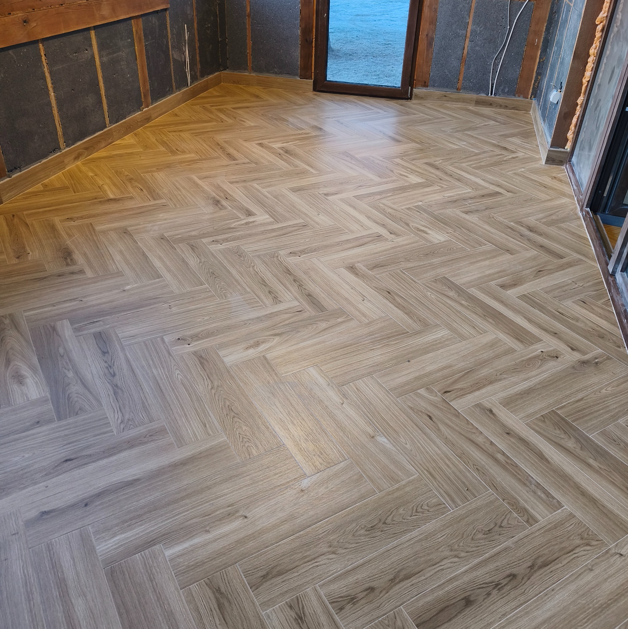 Ułożona podłoga z płytek imitujących drewno w jodełkę, widok z góry. Płytki w odcieniach beżu i brązu. Ściany w trakcie wykończenia, widoczne elementy konstrukcyjne.