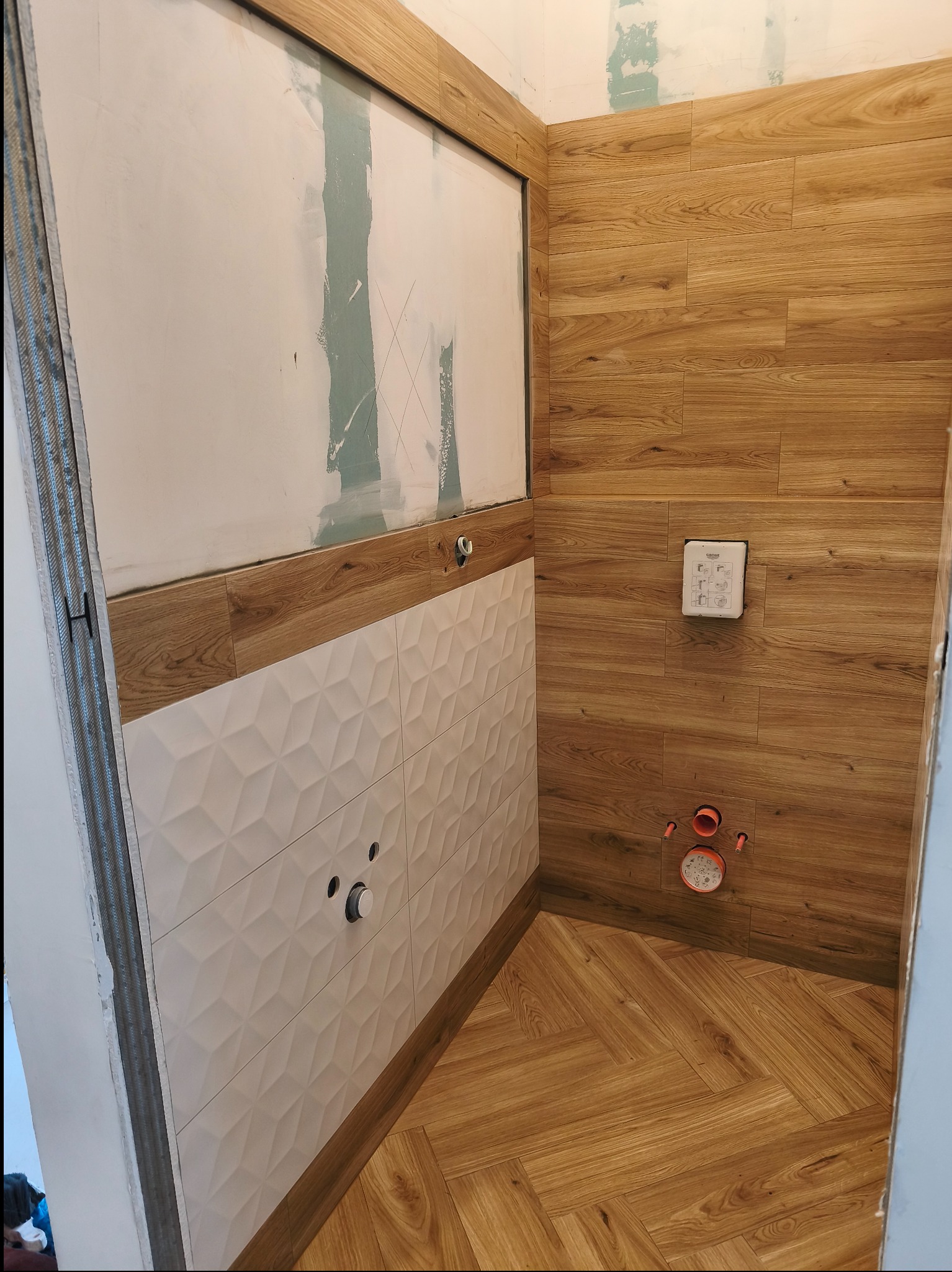 Łazienka w trakcie wykończenia: drewnopodobne płytki na ścianach i podłodze ułożone w jodełkę, białe płytki 3D na ścianie, widoczne instalacje. Częściowo wykończona.