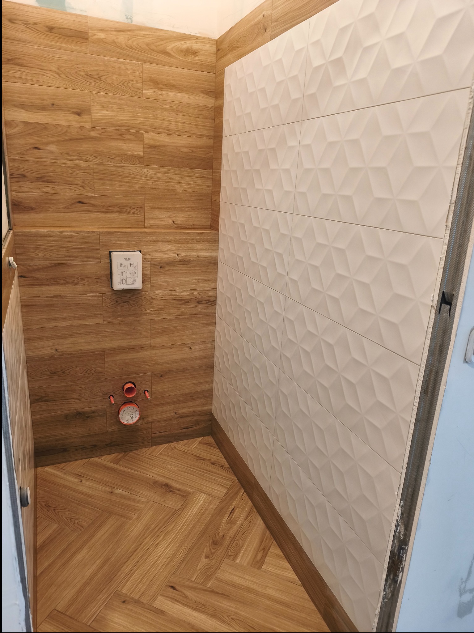 Wykończona kabina prysznicowa z drewnopodobnymi płytkami na ścianach i podłodze ułożonymi w jodełkę oraz białą ścianą z trójwymiarowych płytek.