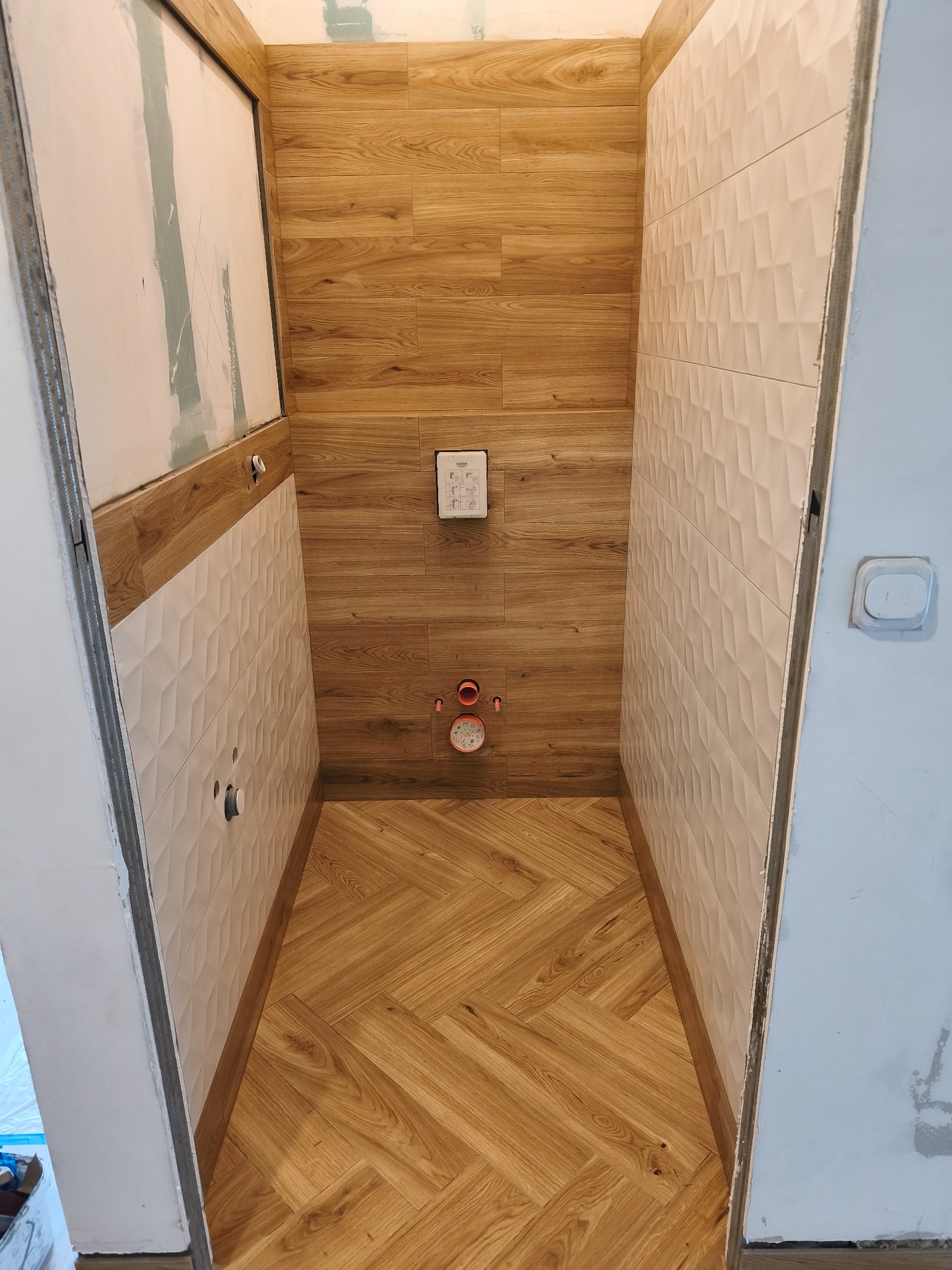 Wnętrze łazienki w trakcie wykańczania: drewnopodobne płytki na ścianach i podłodze ułożone w jodełkę, białe płytki 3D na ścianie, widoczne elementy instalacji.