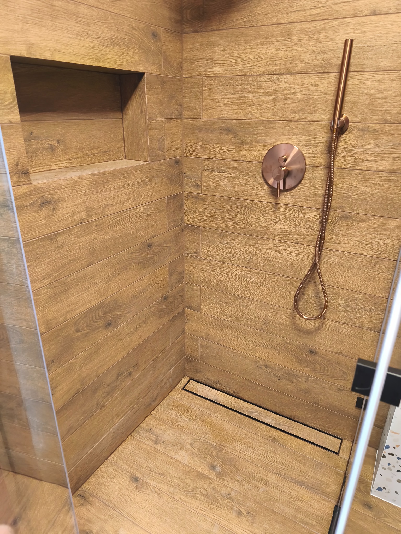 Nowoczesna kabina prysznicowa z drewnopodobnymi płytkami, miedzianą armaturą i odpływem liniowym. Wnęka w ścianie wykończona tym samym materiałem.