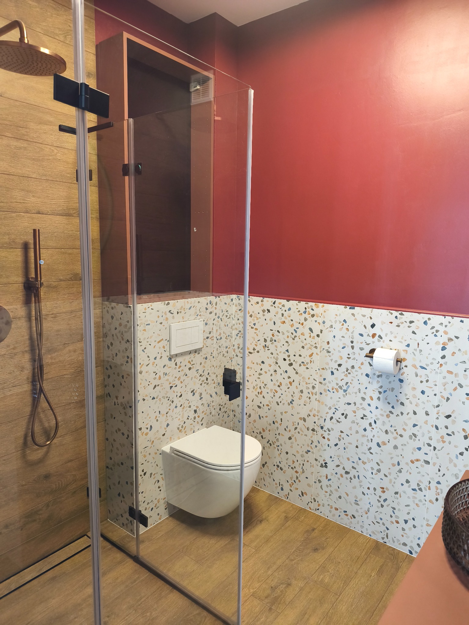 Nowoczesna łazienka z kabiną prysznicową typu walk-in, toaletą podwieszaną i płytkami lastryko na ścianie. Drewnopodobne płytki na podłodze i czerwona ściana.