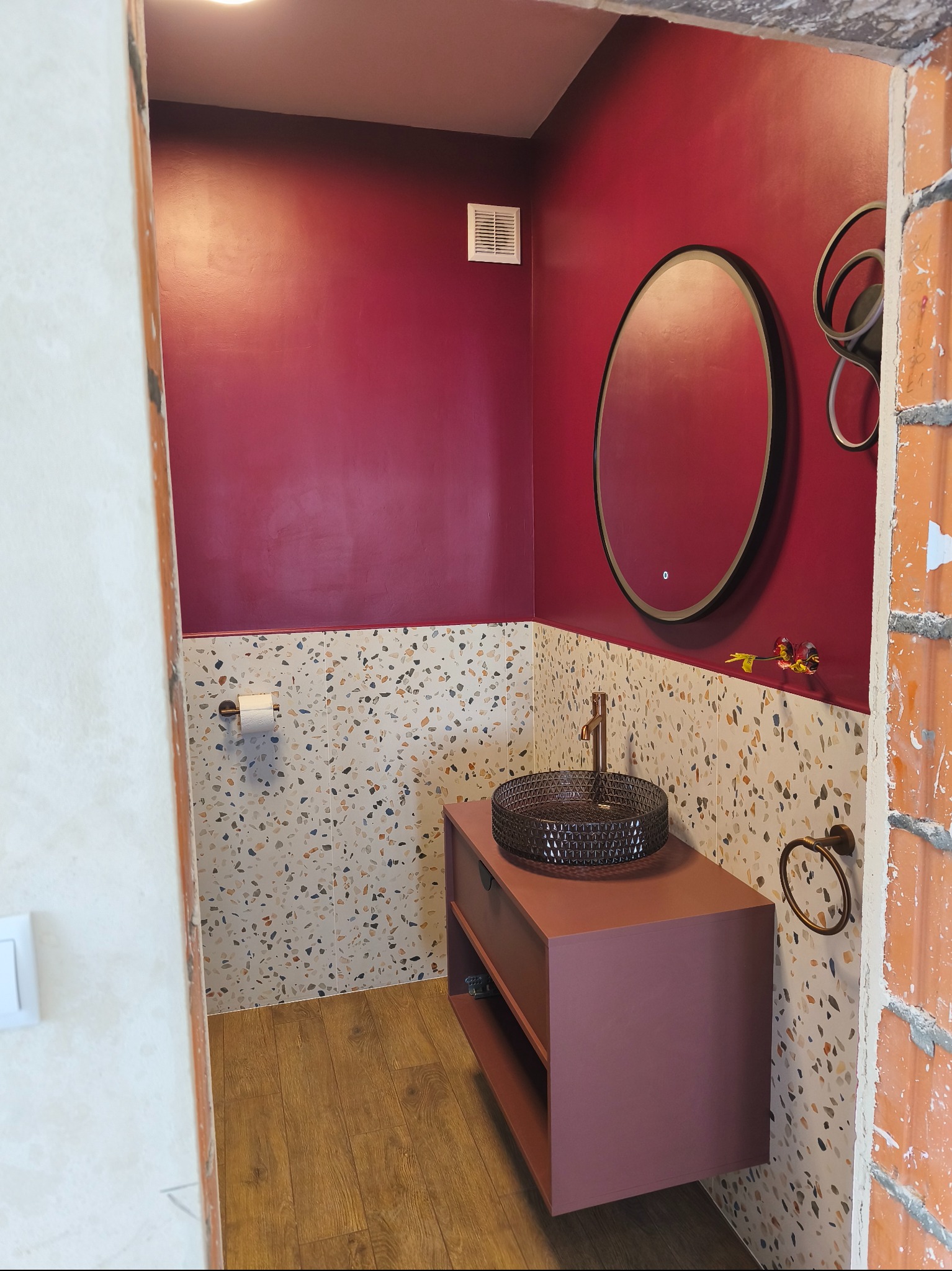 Nowoczesna łazienka w trakcie wykańczania: bordowe ściany, mozaika na ścianie, umywalka na szafce w kolorze bordo, okrągłe lustro z podświetleniem.