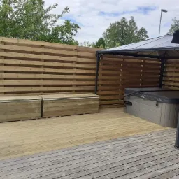 Drewniany taras z jacuzzi pod zadaszeniem i drewnianym ogrodzeniem horyzontalnym, dwa drewniane pojemniki na tarasie, częściowo przykryty grill, widok na drzewa w tle.