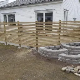 Nowoczesne ogrodzenie z poziomych desek, betonowe słupki i trzy okrągłe konstrukcje z betonowych bloczków na trawniku przed białym domem z szarą dachówką.