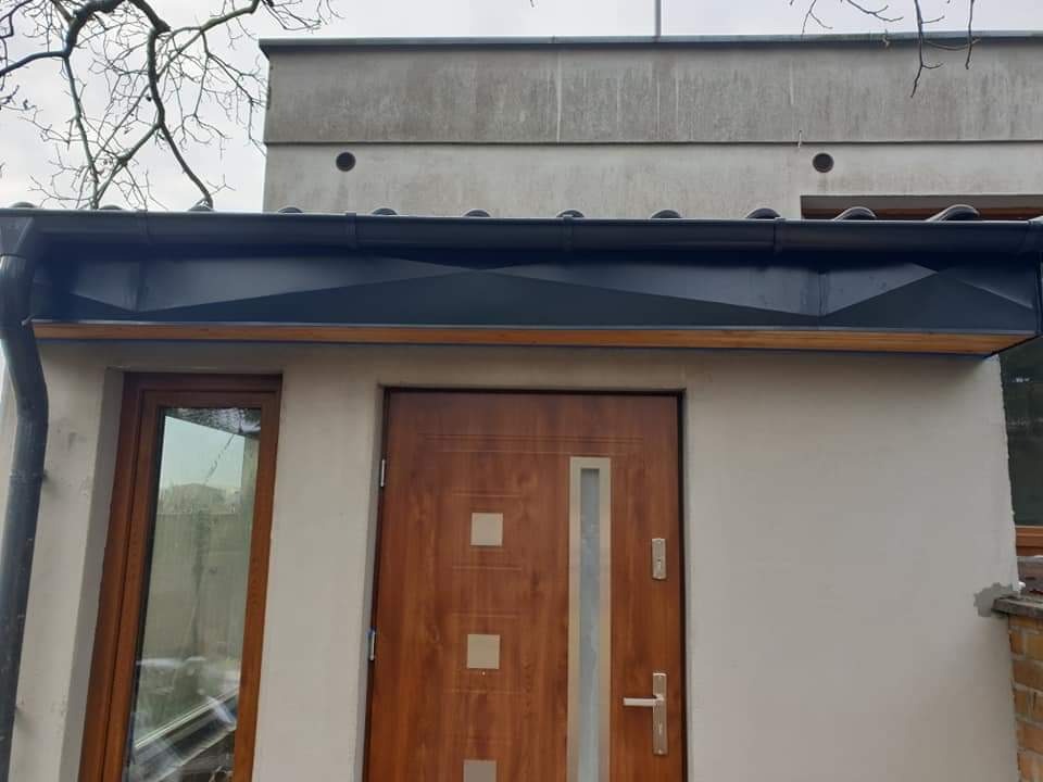 Wejście do budynku z drewnianymi drzwiami i oknem, osłonięte czarnym, metalowym daszkiem z widoczną rynną spustową, na tle szarej elewacji i nagich gałęzi drzew
