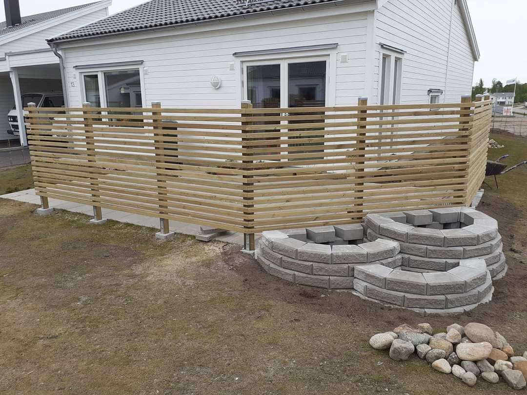 Nowoczesne ogrodzenie z poziomych desek, betonowe słupki i trzy okrągłe konstrukcje z betonowych bloczków na trawniku przed białym domem z szarą dachówką.