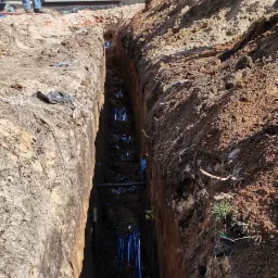 Głęboki wykop w ziemi z widocznymi niebieskimi rurami instalacji, fragment budynku w tle, osoba obserwująca prace.