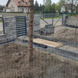 Budowa podjazdu z kostki brukowej: widoczne obrzeża, piasek, ułożona geowłóknina, palety z kostką, częściowo zamontowane ogrodzenie panelowe z betonowych bloczków, teren przygotowany pod dalsze prace.