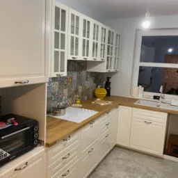 Jasna kuchnia z białymi szafkami, drewnianym blatem, mozaiką nad blatem i piekarnikiem w zabudowie, okno z widokiem na remontowane pomieszczenie.