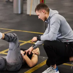 Kobieta w legginsach ćwiczy pod okiem mężczyzny w bluzie z kapturem, leżąc na macie w siłowni. Mężczyzna koryguje pozycję kobiety.