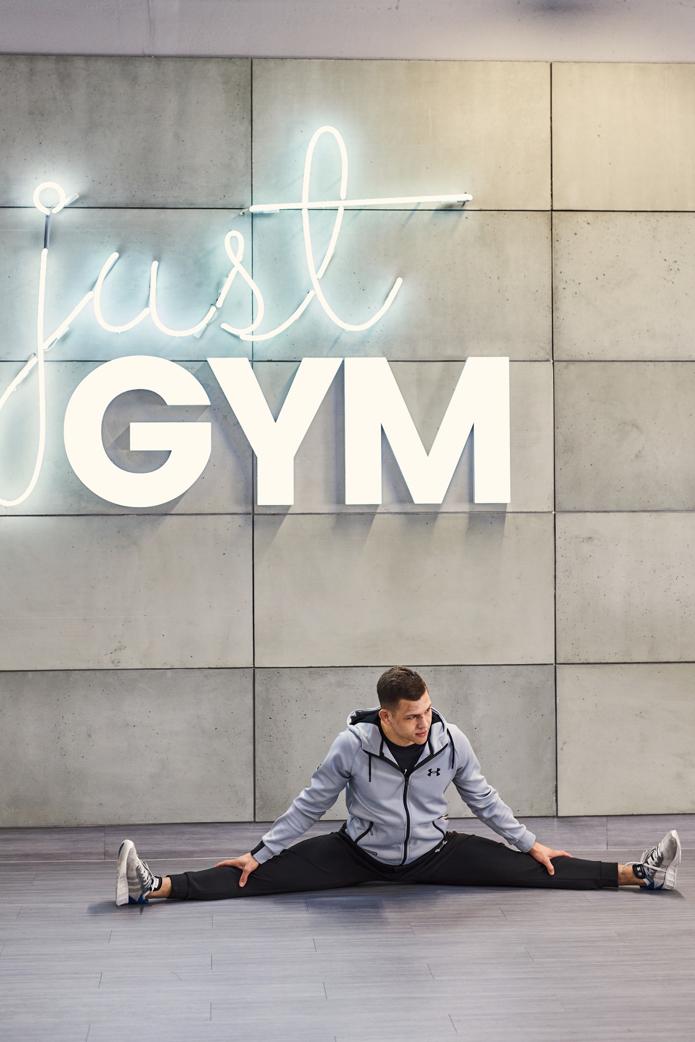 Mężczyzna w szarej bluzie z kapturem wykonuje szpagat na podłodze w siłowni, na ścianie neonowy napis 'just GYM'.