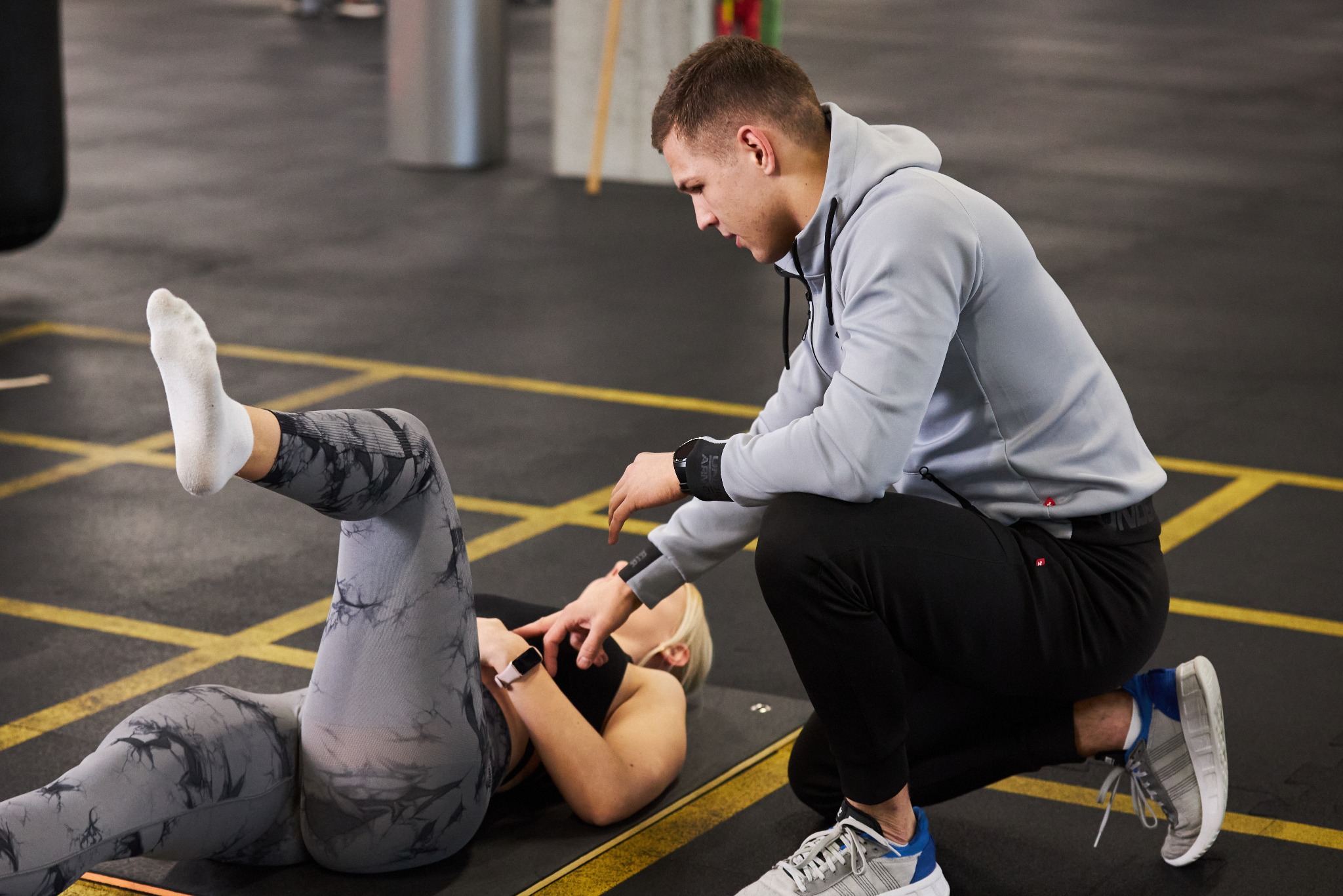 Kobieta w legginsach ćwiczy pod okiem mężczyzny w bluzie z kapturem, leżąc na macie w siłowni. Mężczyzna koryguje pozycję kobiety.