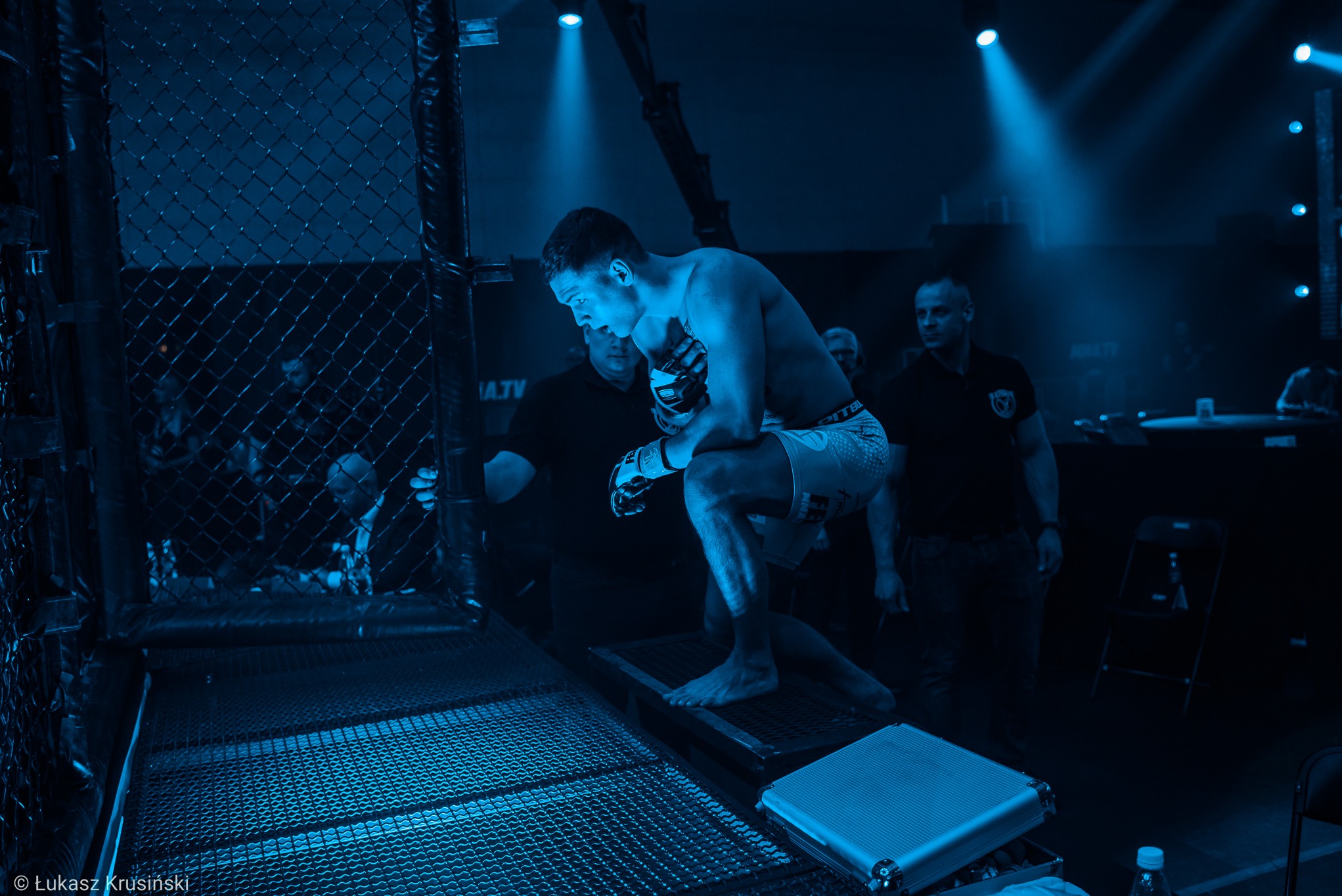 Zawodnik MMA wchodzi do klatki, przygotowując się do walki w niebieskim oświetleniu, widoczny fragment klatki, publiczność i obsługa techniczna w tle.