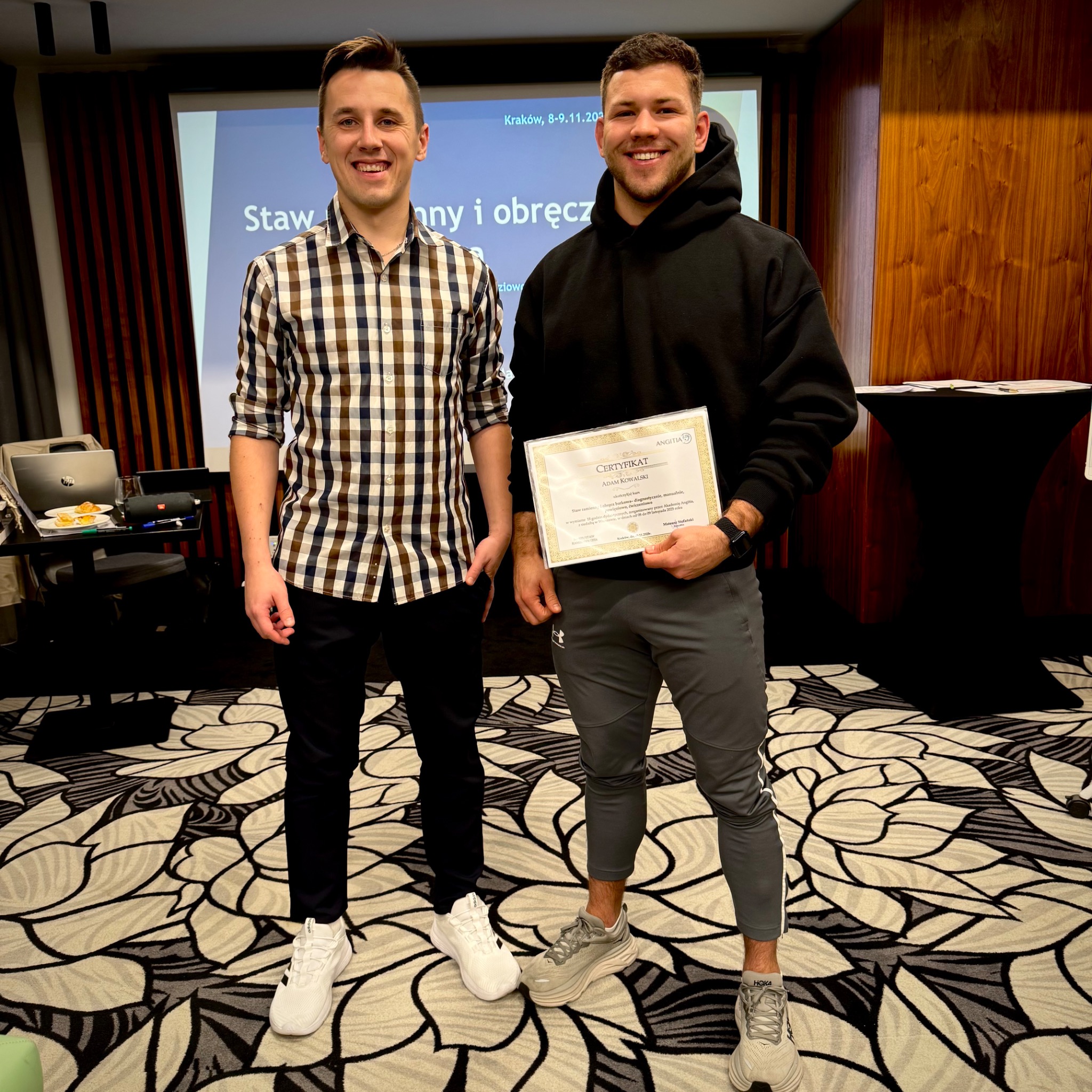 Dwóch uśmiechniętych mężczyzn prezentuje certyfikat ukończenia kursu z zakresu diagnostyki i terapii stawu ramiennego na tle sali konferencyjnej.