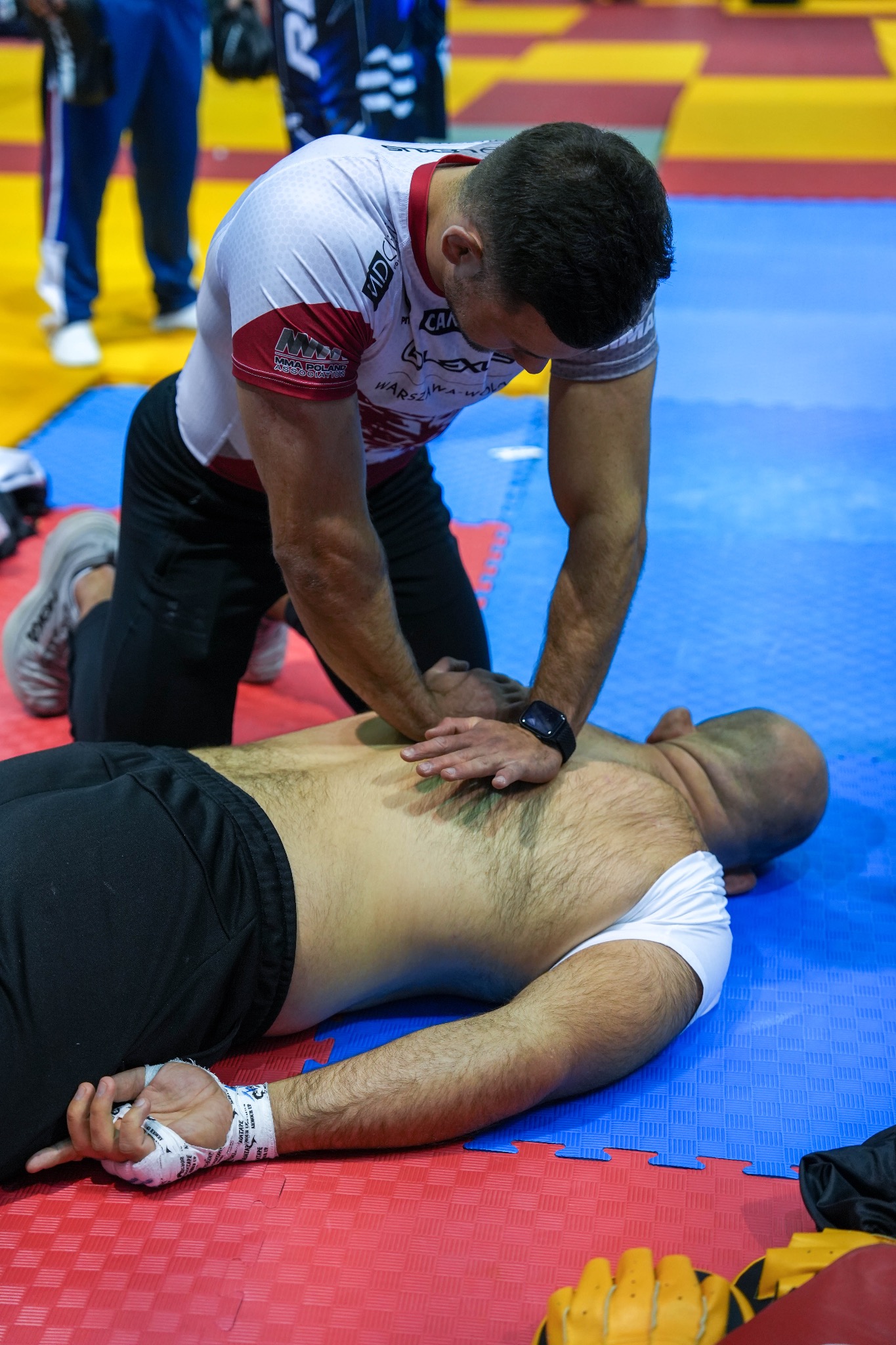 Mężczyzna w białej koszulce uciska klatkę piersiową leżącego mężczyzny na czerwono-niebieskiej macie. Widoczne bandaże na dłoni i sprzęt sportowy w tle.