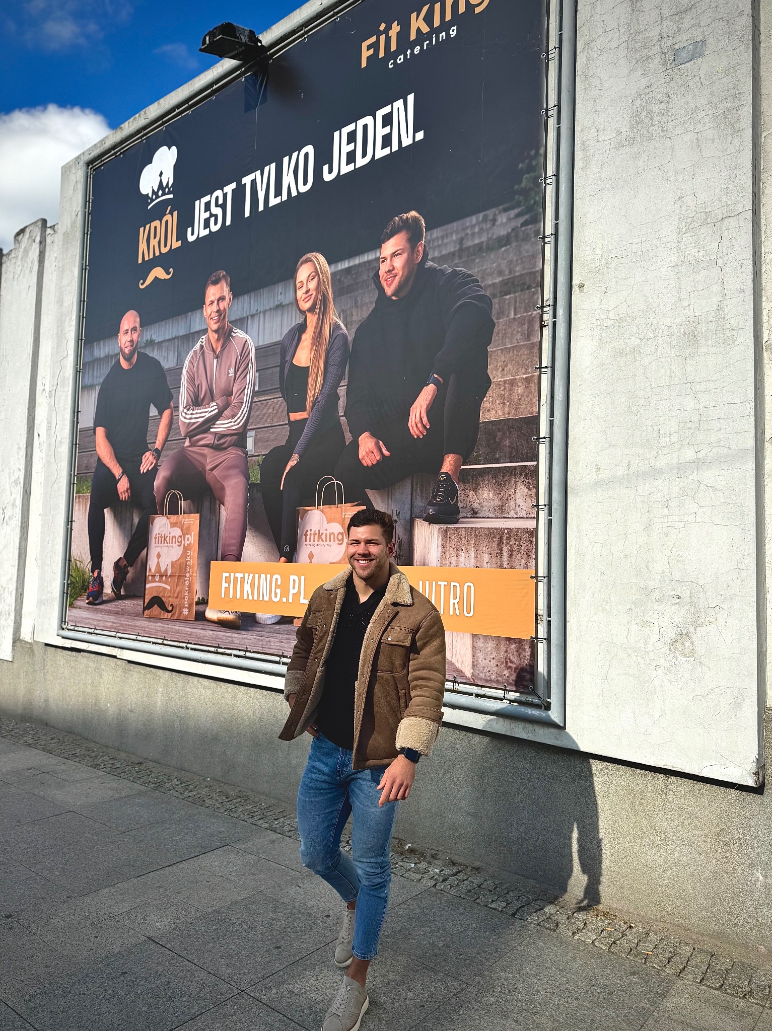 Uśmiechnięty mężczyzna w jeansach i kurtce pozuje na tle billboardu Fit King Catering z wizerunkami sportowców i hasłem 'Król jest tylko jeden'.