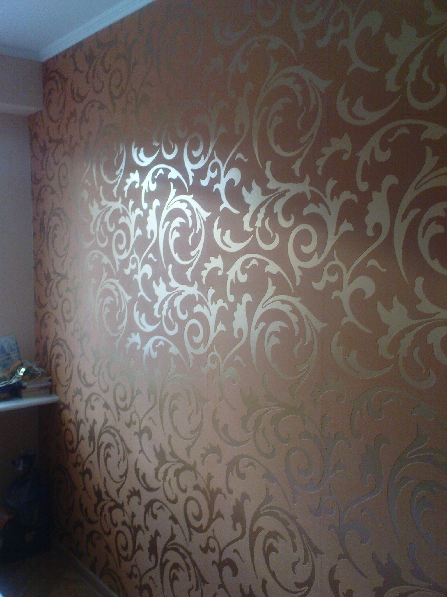 Ściana pokryta tapetą w odcieniach brązu z delikatnym, metalicznym wzorem roślinnym w stylu secesyjnym, widoczna z bliska, oświetlona z lewej strony.