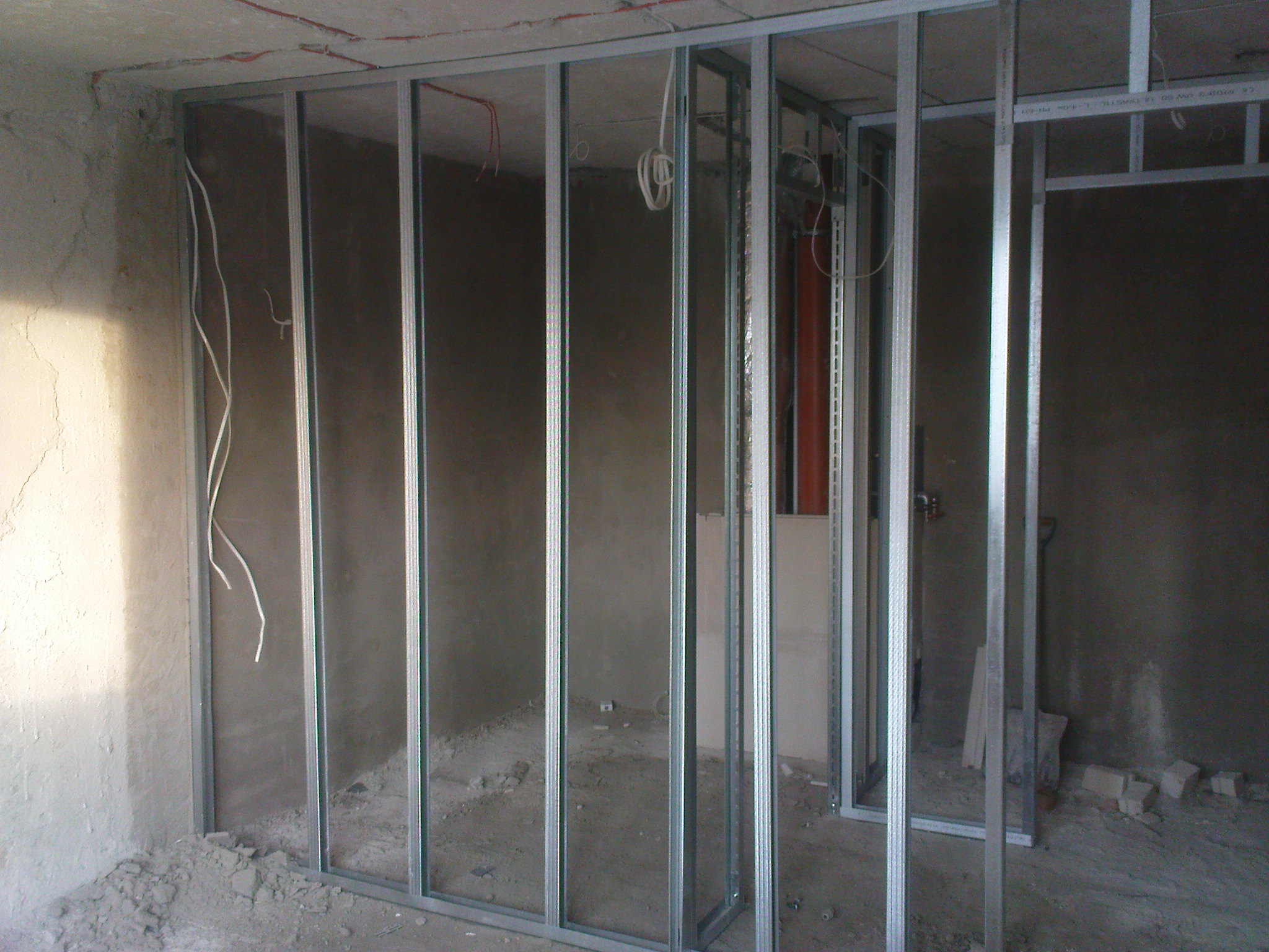 Stelaż z profili metalowych do ścian działowych w surowym stanie, widoczne kable elektryczne wystające ze ściany, podłoże z resztkami gruzu.