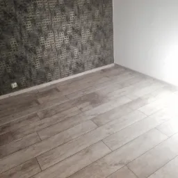 Ułożone panele podłogowe imitujące drewno w pomieszczeniu z tapetą o wzorze geometrycznym i białą ścianą.