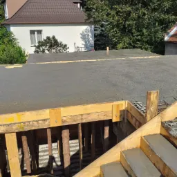 Zalewanie betonem stropu i schodów w nowo powstającym budynku z drewnianym szalunkiem, widoczne zbrojenie i dom w tle.