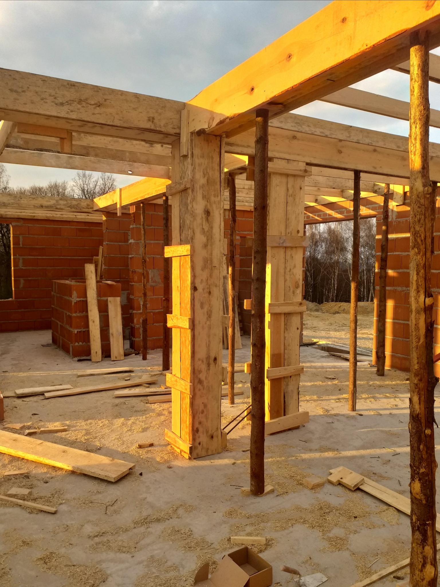 Wnętrze surowego domu w trakcie budowy, widoczne ceglane ściany, drewniane stemple i szalunki podtrzymujące strop, resztki drewna na podłodze, ciepłe światło słoneczne.
