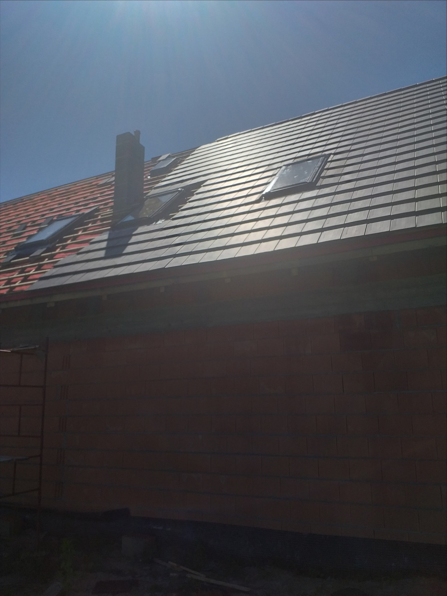 Częściowo pokryty dach z dachówką ceramiczną w kolorze grafitowym, widoczne okna dachowe i komin, fragment ściany budynku z czerwonej cegły.