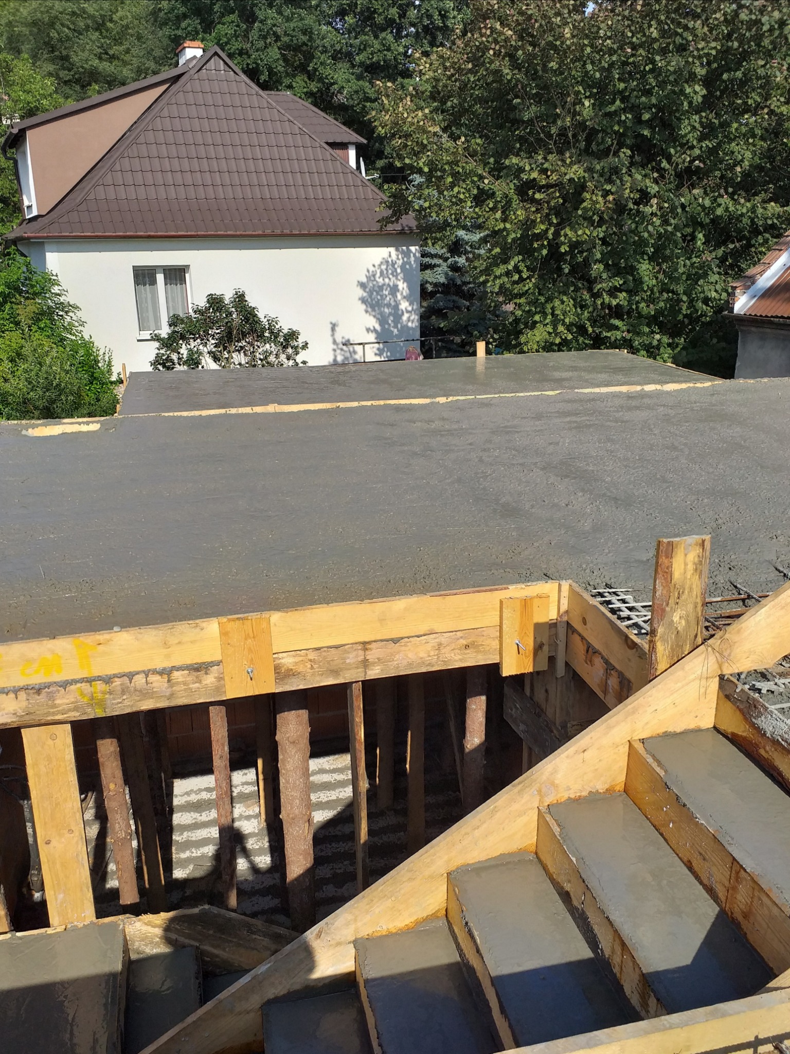 Zalewanie betonem stropu i schodów w nowo powstającym budynku z drewnianym szalunkiem, widoczne zbrojenie i dom w tle.