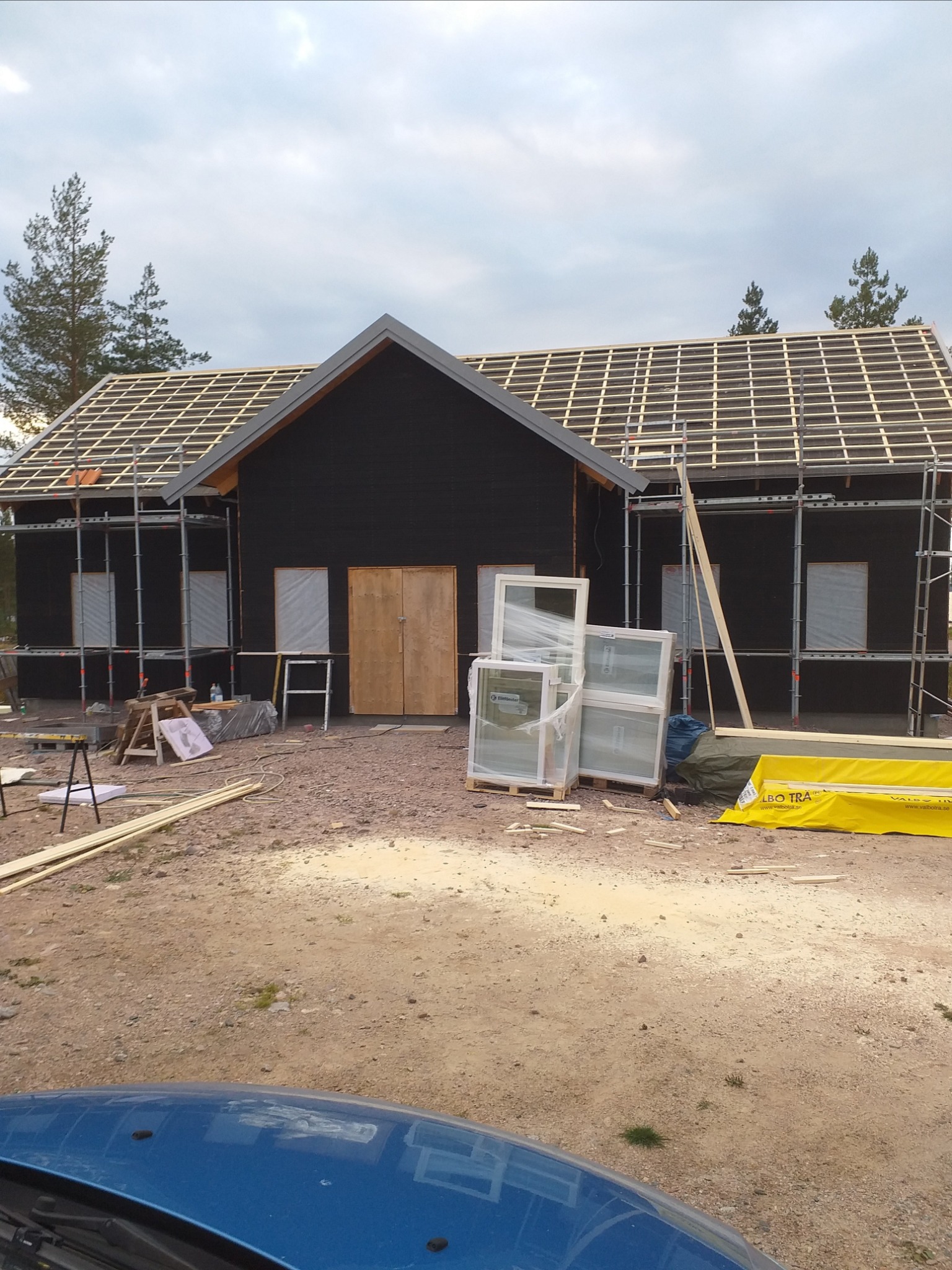 Czarny dom w trakcie budowy, z drewnianą konstrukcją dachu, rusztowaniami, nowymi oknami w folii i niezagospodarowanym terenem.