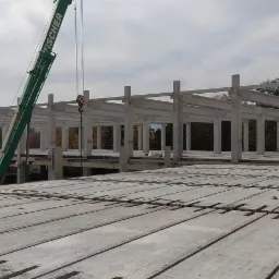 Konstrukcja hali w trakcie budowy: widok z góry na ułożone betonowe płyty stropowe, w tle szkielet hali z prefabrykatów betonowych i zielony dźwig marki FISCHER.