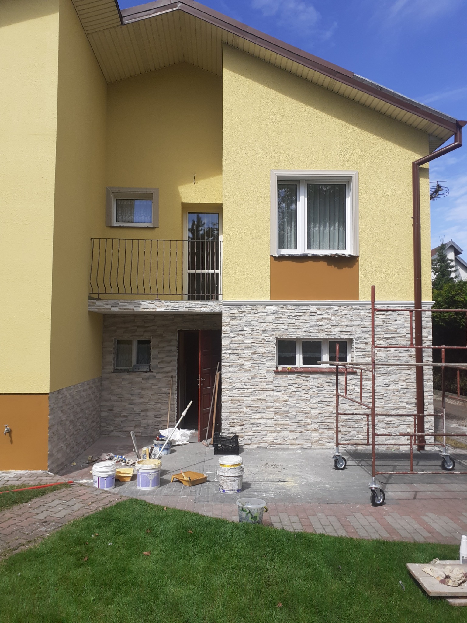 Żółta elewacja domu dwukondygnacyjnego z dekoracyjnym panelem z kamienia, widoczne rusztowanie na kółkach, otwarte drzwi wejściowe, oraz puszki z farbą i narzędzia malarskie na brukowanym podłożu.