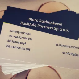 Stos wizytówek Biura Rachunkowego KaskAda Partners Sp. z o.o. na tle drewnianych podkładek, z widocznymi danymi kontaktowymi Katarzyny Fuchs i Adrianny Czyż oraz adresem w Tychach.