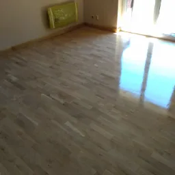 Świeżo położona, jasna podłoga parkietowa z widocznym odblaskiem światła z okna balkonowego, listwy przypodłogowe w kolorze drewna, żółta folia zabezpieczająca na ścianie.