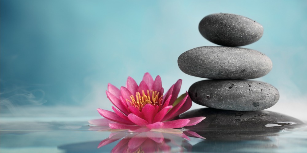 Trzy ułożone jeden na drugim szare kamienie obok różowego kwiatu lotosu odbijającego się w wodzie na tle błękitnego nieba.