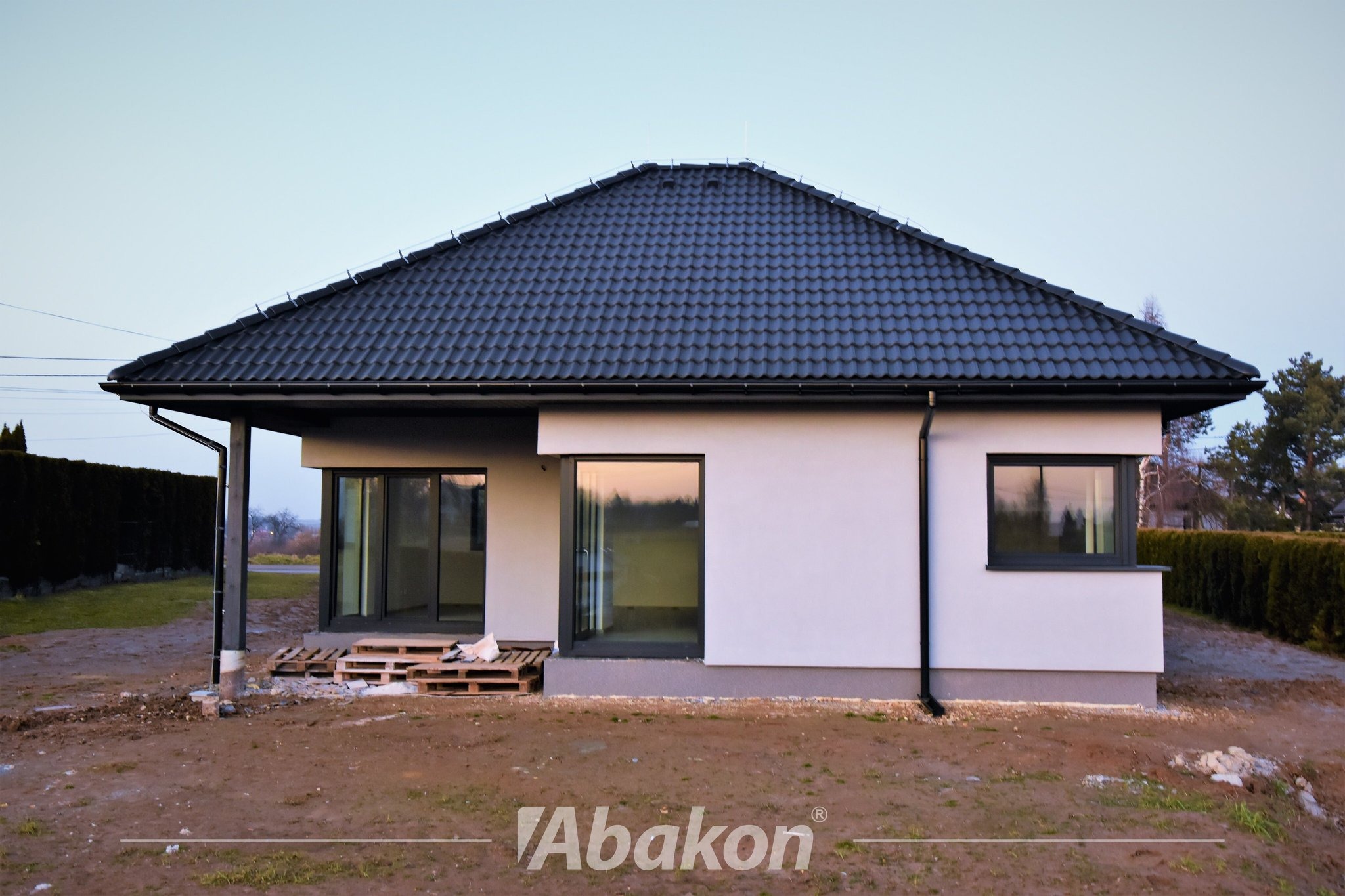 Nowoczesny, parterowy dom z ciemną dachówką i szarą stolarką okienną w trakcie budowy, widoczne palety i przygotowany teren wokół budynku.