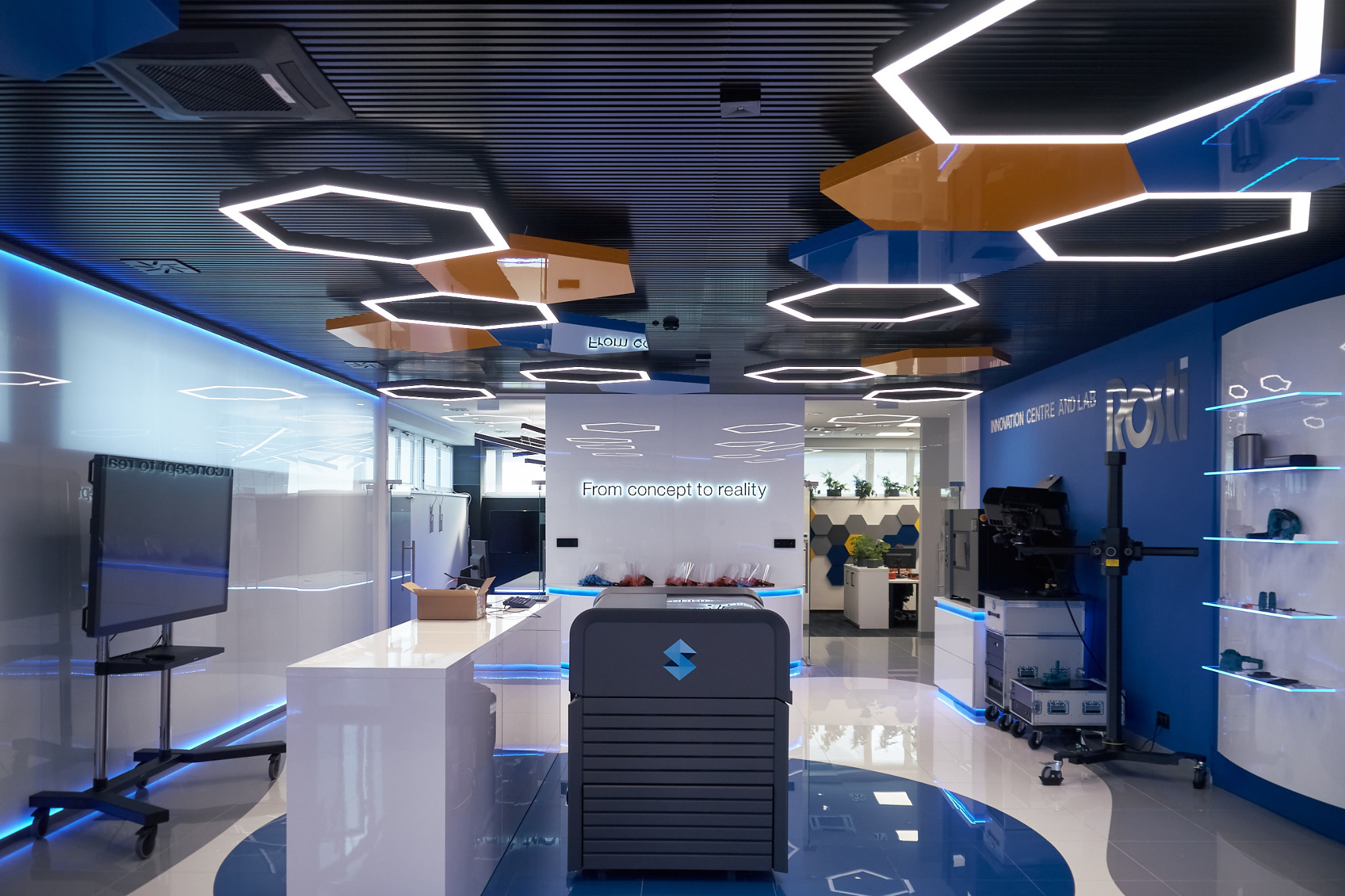 Nowoczesne wnętrze biurowe w Białymstoku z heksagonalnym oświetleniem sufitowym, białymi meblami z podświetleniem LED, niebieskimi akcentami kolorystycznymi i napisem 'From concept to reality'.