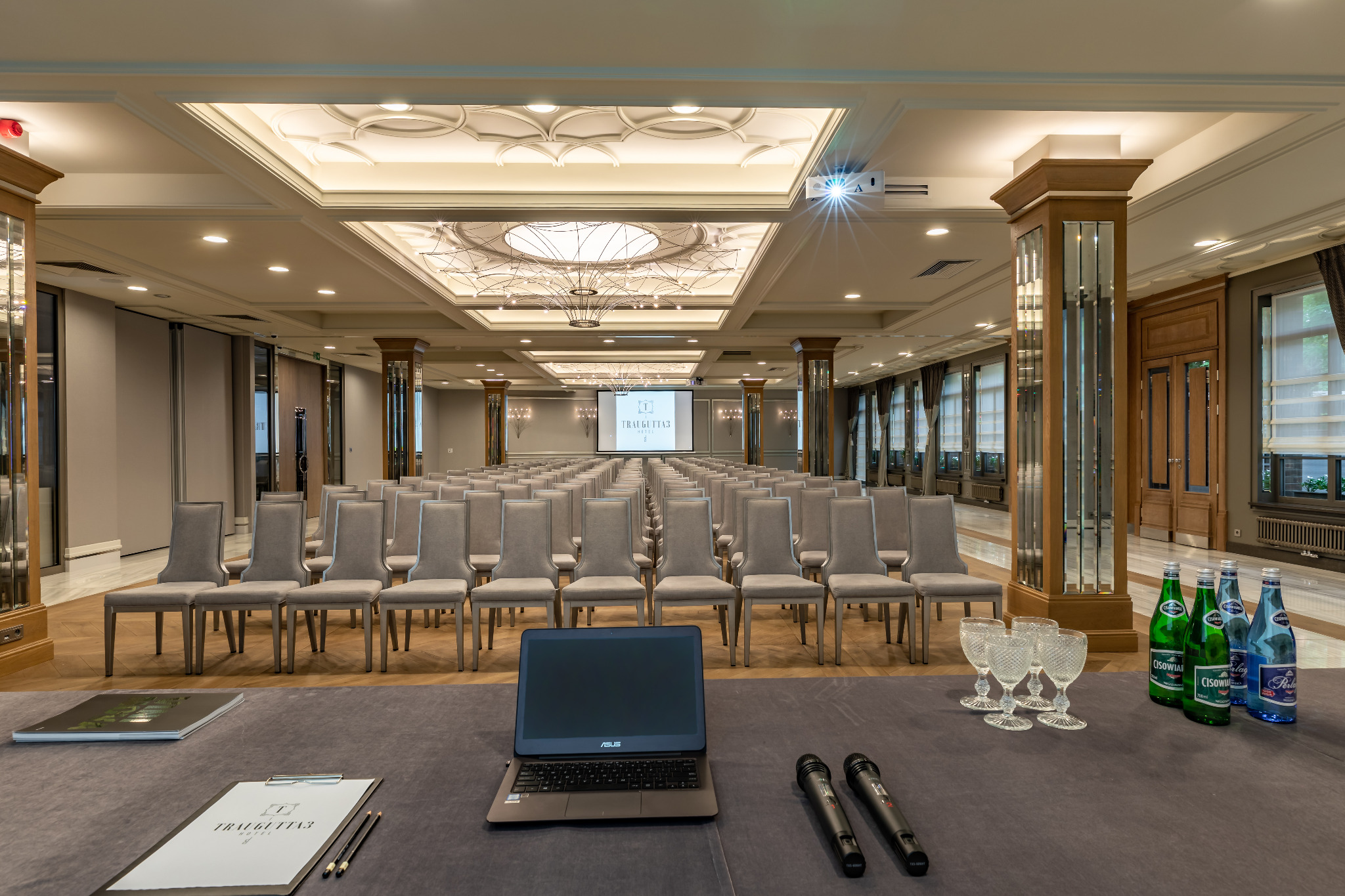 Sala konferencyjna Hotelu Traugutta3 w Białymstoku przygotowana na spotkanie: rzędy krzeseł, laptop, mikrofony, woda mineralna i kieliszki na pierwszym planie, projektor sufitowy i ekran z logo...