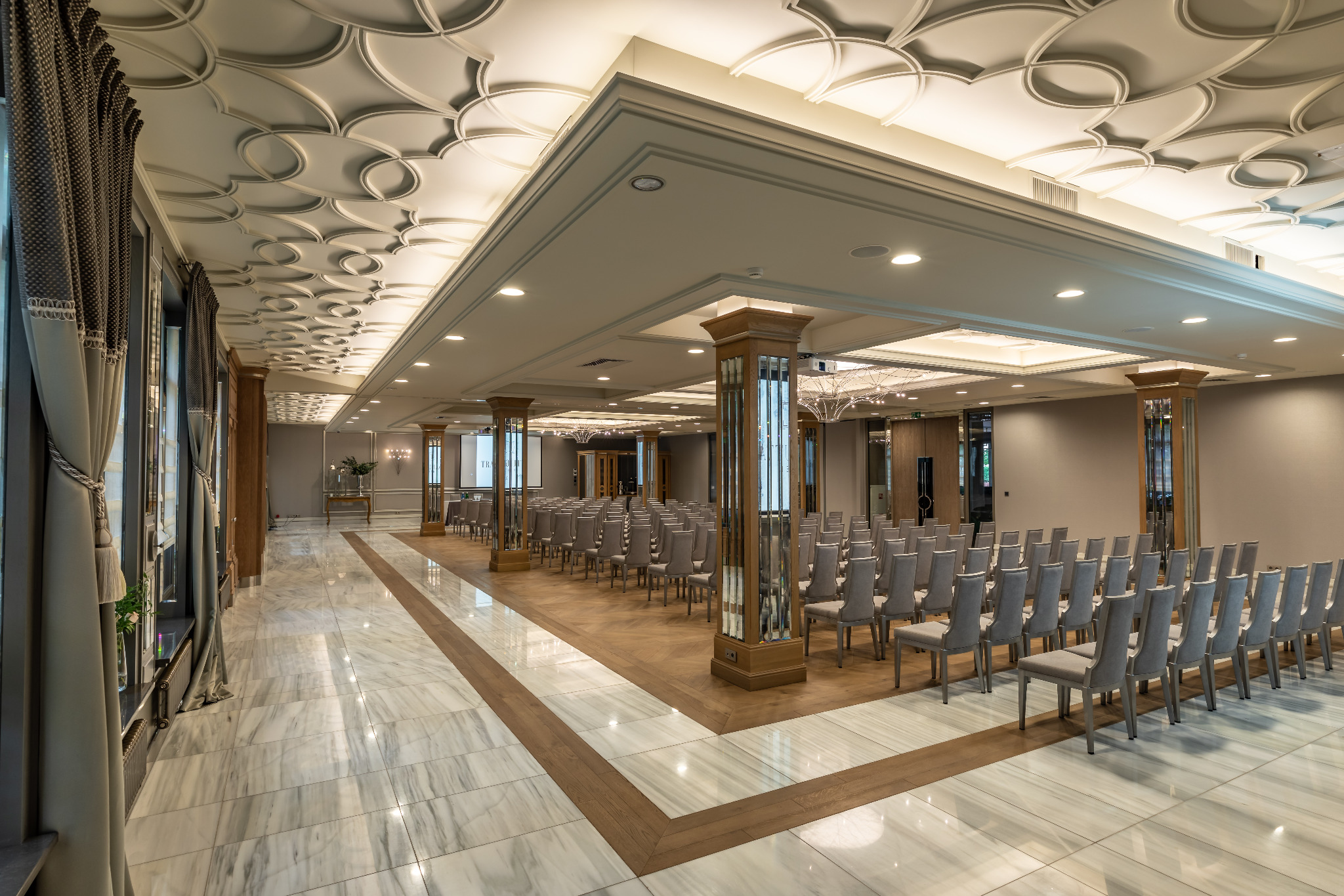Sala konferencyjna w Hotelu Traugutta3 w Białymstoku z rzędami szarych krzeseł ustawionych na drewnianym podeście, marmurowa podłoga i kolumny z lustrami, dekoracyjny sufit z oświetleniem.