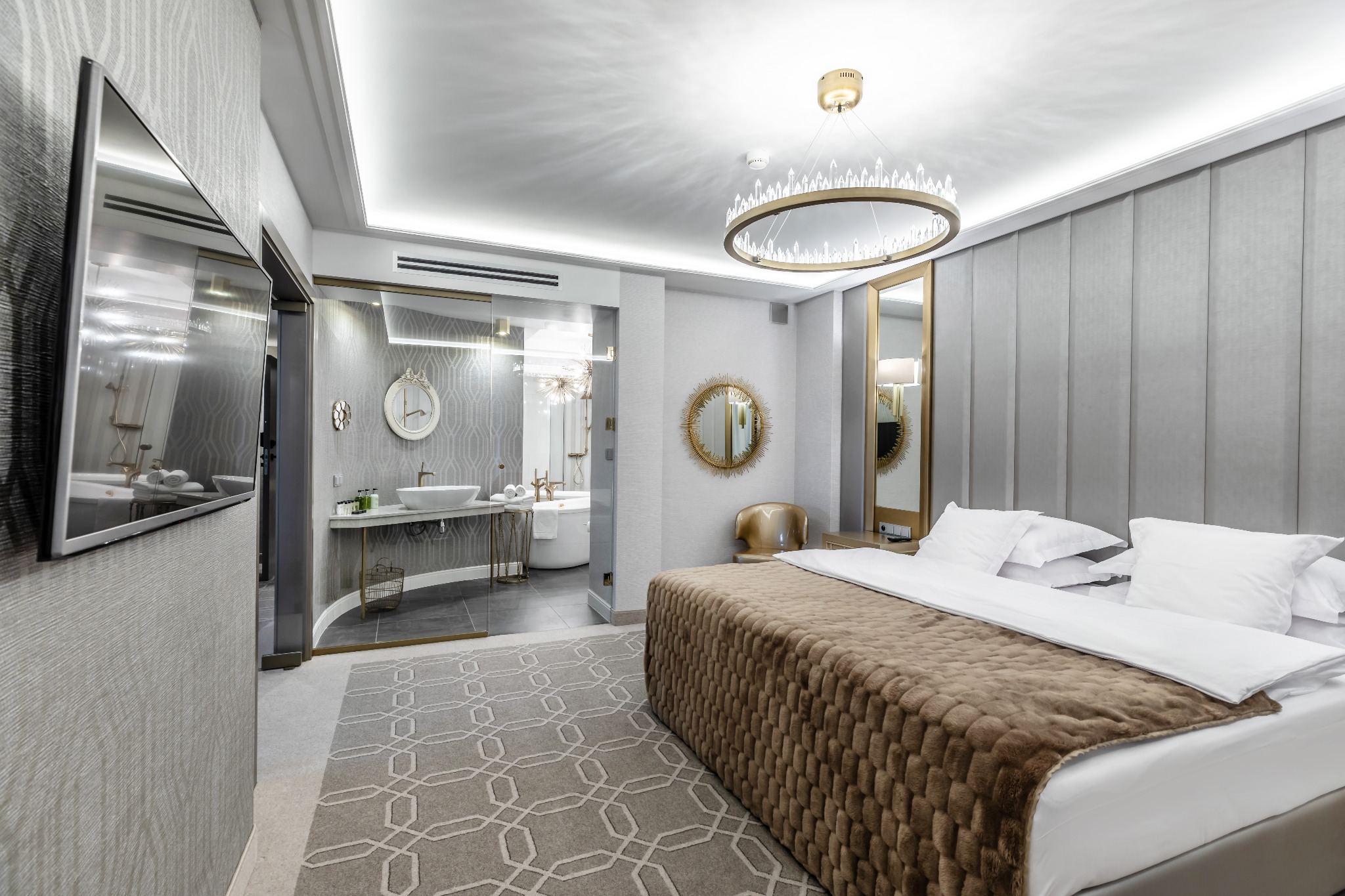 Nowoczesny pokój hotelowy z widokiem na łazienkę przez szklaną ścianę, okrągły żyrandol, neutralna kolorystyka, telewizor na ścianie, wzorzysty dywan.