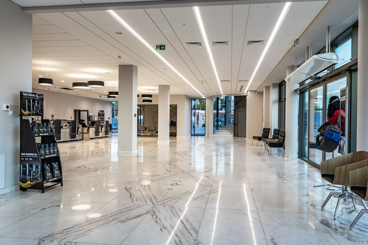 Przestronne, jasne wnętrze salonu samochodowego z marmurową podłogą, oświetleniem liniowym LED, czarnymi lampami sufitowymi i ekspozytorem oleju silnikowego MAN Genuine Oil przy ścianie.