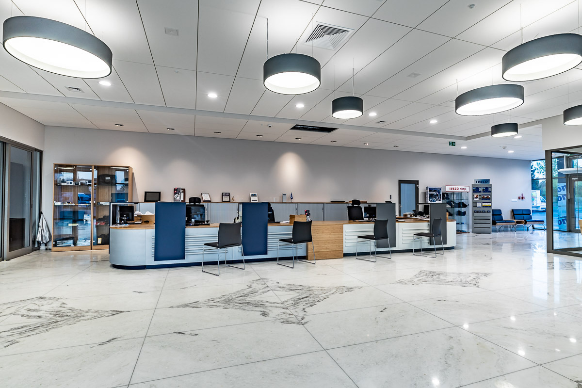 Wnętrze salonu samochodowego Targor-Truck w Rzekuń z nowoczesnym oświetleniem w postaci okrągłych lamp sufitowych, marmurową podłogą i ladą recepcyjną z elementami drewna i niebieskiego akcentu.