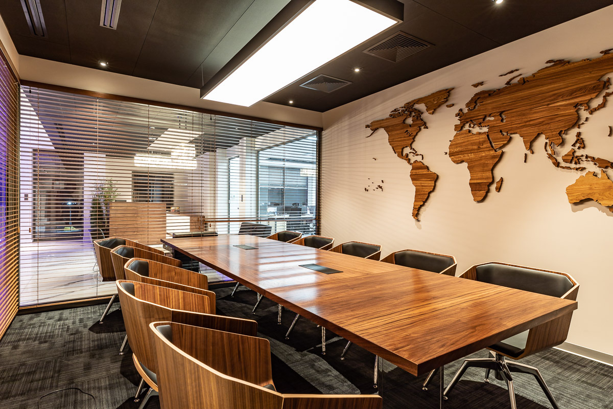Nowoczesna sala konferencyjna z długim drewnianym stołem, krzesłami z drewnianymi oparciami i czarnymi siedziskami, drewnianą mapą świata na ścianie oraz oknem z żaluzjami.